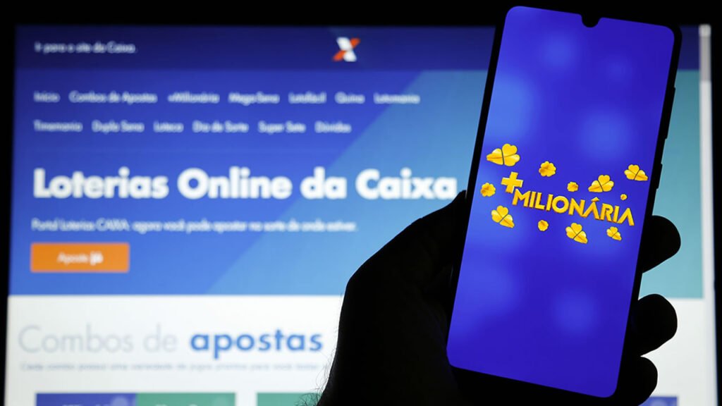 Mão segurando celular, atrás tela do computador no site da Loterias CAIXA