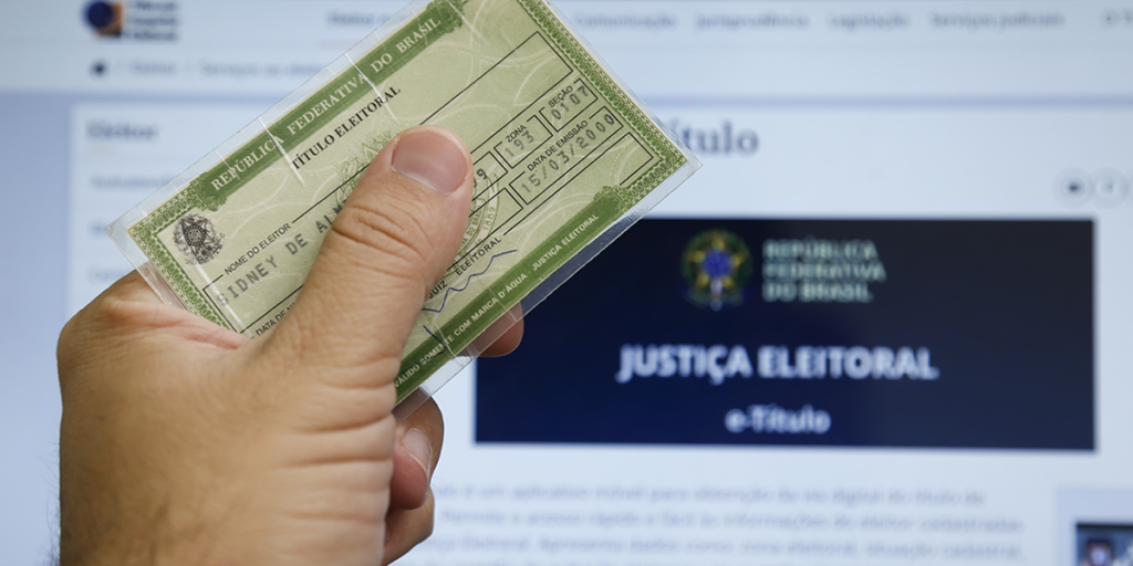 Foto exibe mão segurando título de eleitor e ao fundo site da Justiça Eleitoral