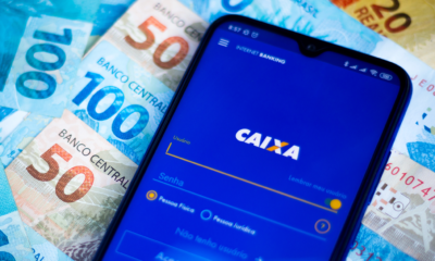 Celular no aplicativo da CAIXA. Ao fundo, notas de dinheiro, reais, brasileiro